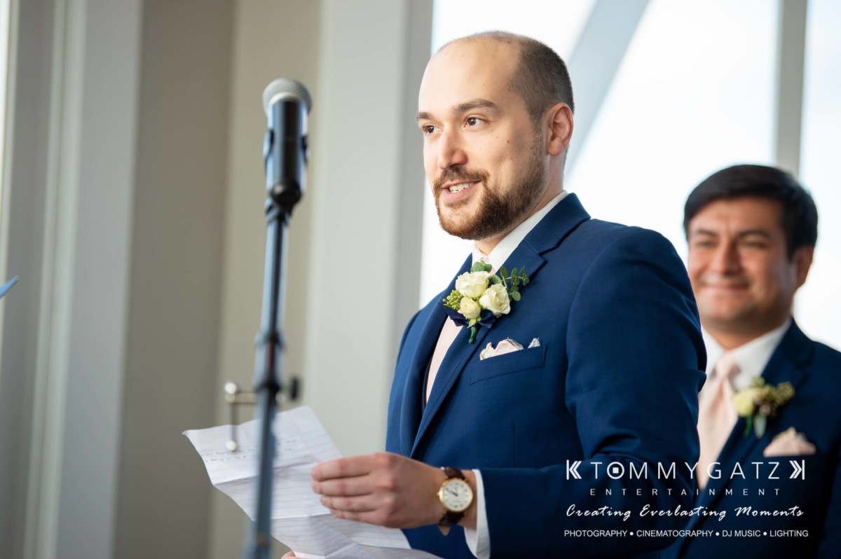 BrittanyDaniel-WEDDING-Highlights-61 - TOMMY GATZ ENTERTAINMENT A man in a suit stands at a microphone holding a piece of paper, while another man in a suit stands smiling in the background.
