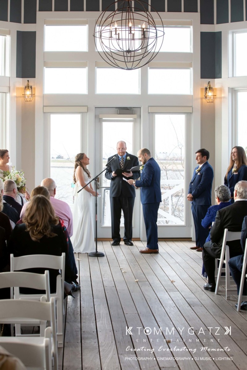 BrittanyDaniel-WEDDING-Highlights-62 - TOMMY GATZ ENTERTAINMENT A wedding ceremony with a bride and groom standing in front of an officiant, surrounded by guests seated on white chairs in a bright room.