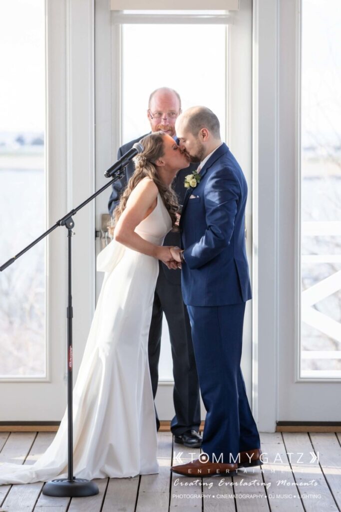 BrittanyDaniel-WEDDING-Highlights-66 - TOMMY GATZ ENTERTAINMENT A couple is sharing a kiss during their wedding ceremony, with a man standing behind them smiling, in front of a bright window.