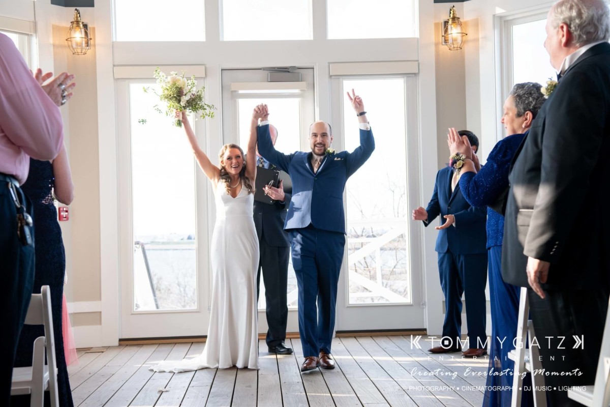 BrittanyDaniel-WEDDING-Highlights-67 - TOMMY GATZ ENTERTAINMENT A newlywed couple celebrates with raised arms indoors, surrounded by clapping guests.