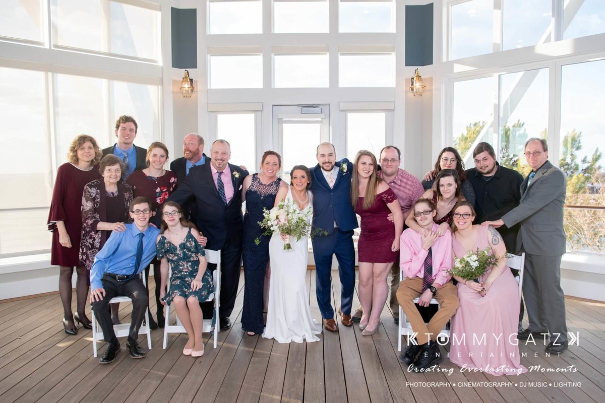 BrittanyDaniel-WEDDING-Highlights-68 - TOMMY GATZ ENTERTAINMENT A wedding group photo with a couple in the center, surrounded by family and friends on a wooden deck. Some hold flowers, and everyone is dressed in formal attire.