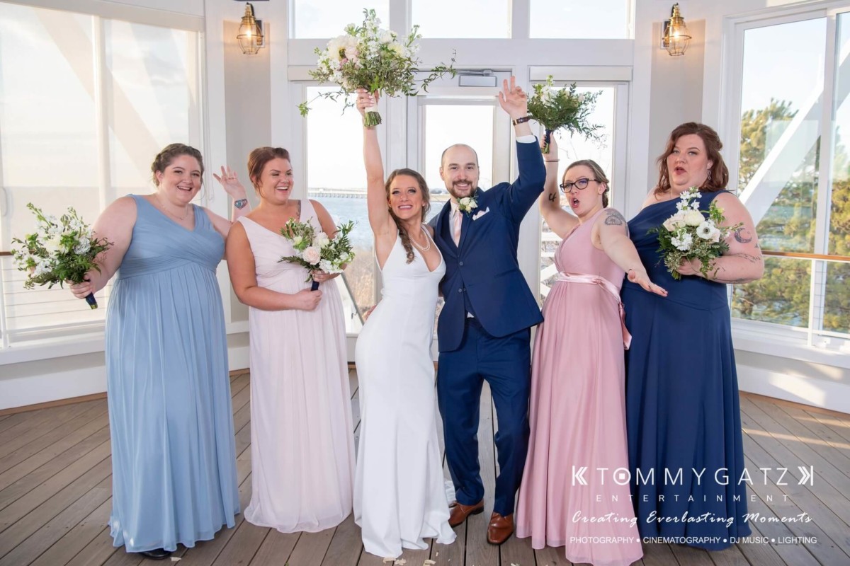 BrittanyDaniel-WEDDING-Highlights-69 - TOMMY GATZ ENTERTAINMENT A wedding couple and four bridesmaids pose cheerfully inside a bright room with bouquets and celebratory gestures.