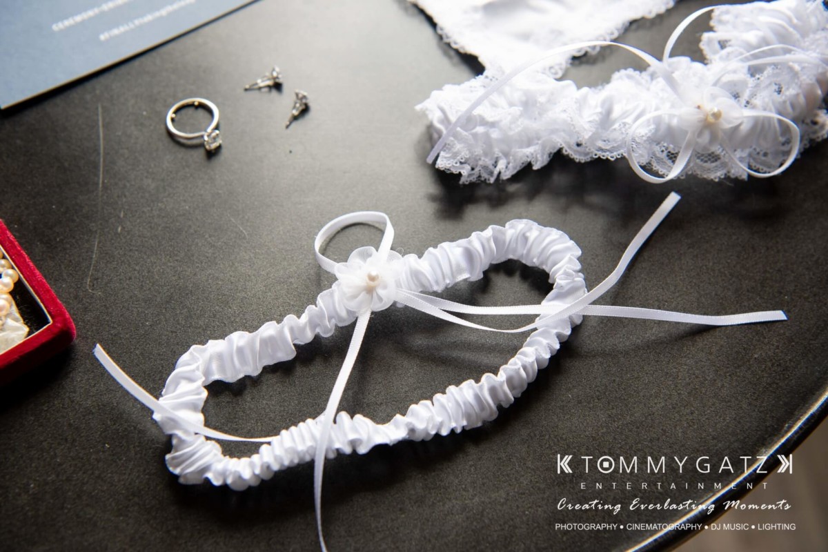 BrittanyDaniel-WEDDING-Highlights-7 - TOMMY GATZ ENTERTAINMENT White bridal garter with ribbons and lace on a table, accompanied by earrings and rings.