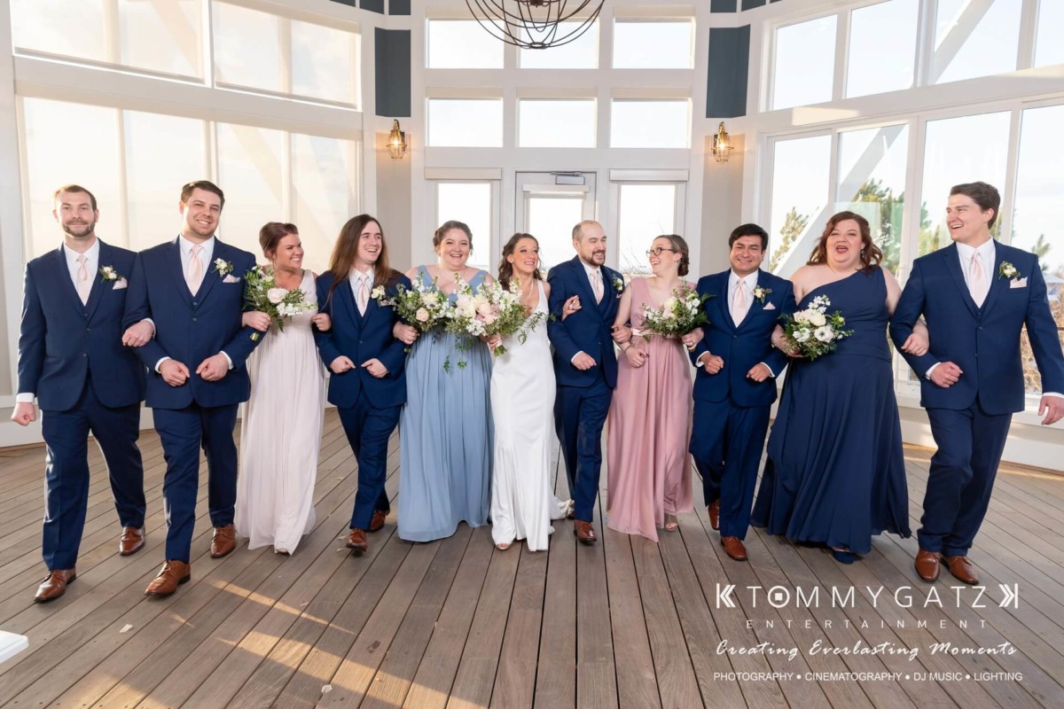BrittanyDaniel-WEDDING-Highlights-70 - TOMMY GATZ ENTERTAINMENT A wedding party walks arm-in-arm indoors. The bridesmaids wear pastel dresses, and the groomsmen wear navy suits with pink ties. The central couple wears a white dress and navy suit.
