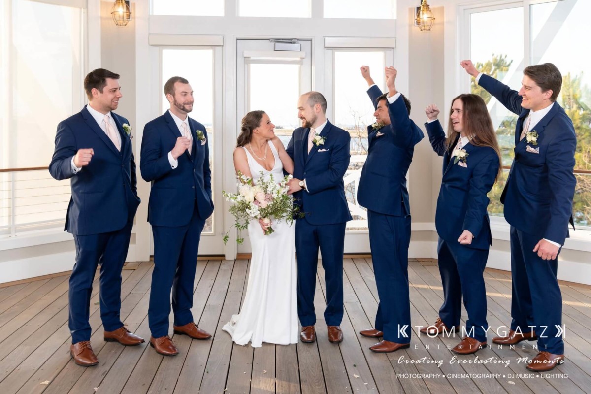 BrittanyDaniel-WEDDING-Highlights-71 - TOMMY GATZ ENTERTAINMENT Bride and groom stand together with groomsmen in blue suits celebrating on a covered porch.