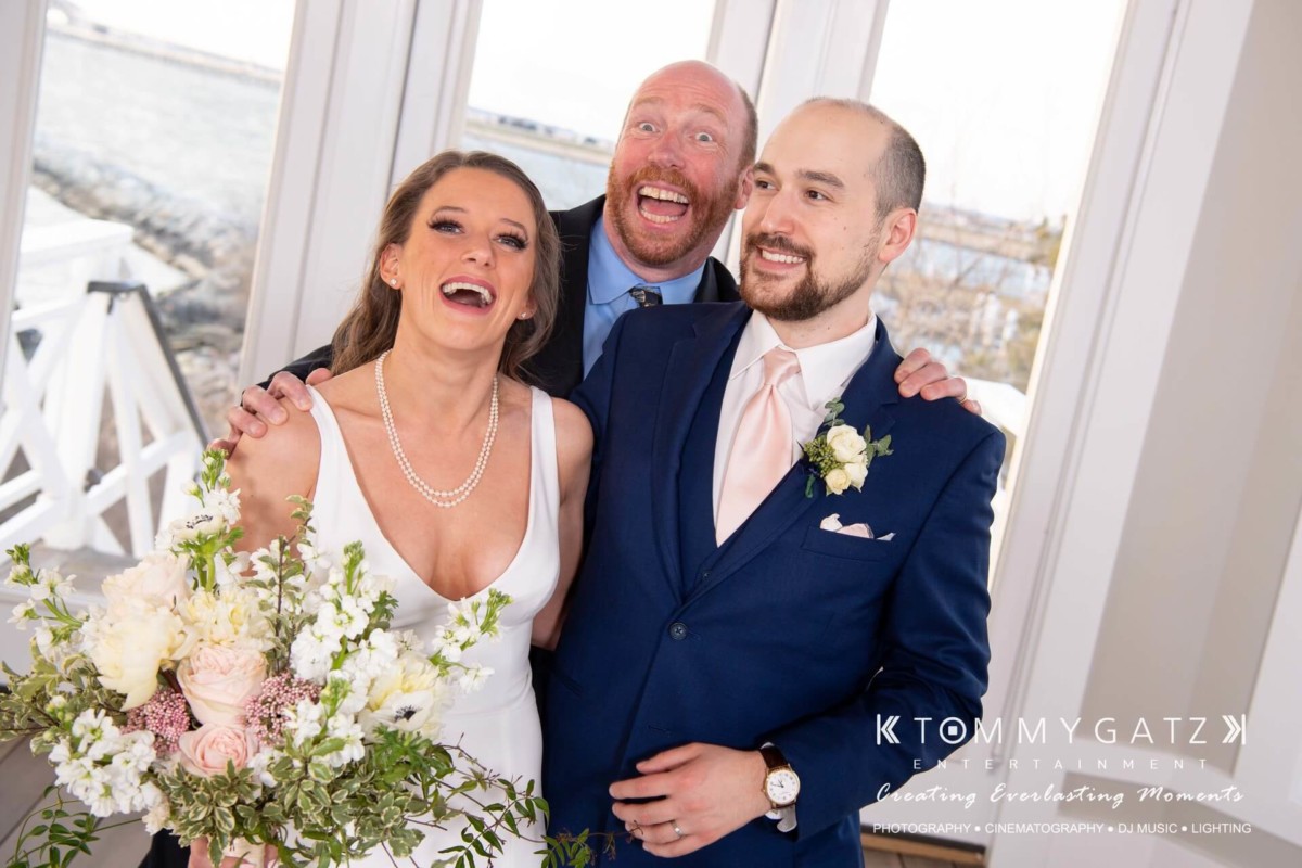BrittanyDaniel-WEDDING-Highlights-72 - TOMMY GATZ ENTERTAINMENT A newlywed couple poses with a smiling man indoors. The bride holds a bouquet of flowers.