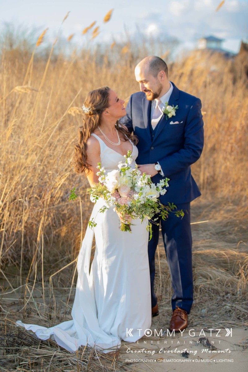 BrittanyDaniel-WEDDING-Highlights-73 - TOMMY GATZ ENTERTAINMENT A couple stands together outdoors. The woman holds a bouquet and wears a white dress, while the man is in a blue suit. They are surrounded by tall grass under a clear sky.