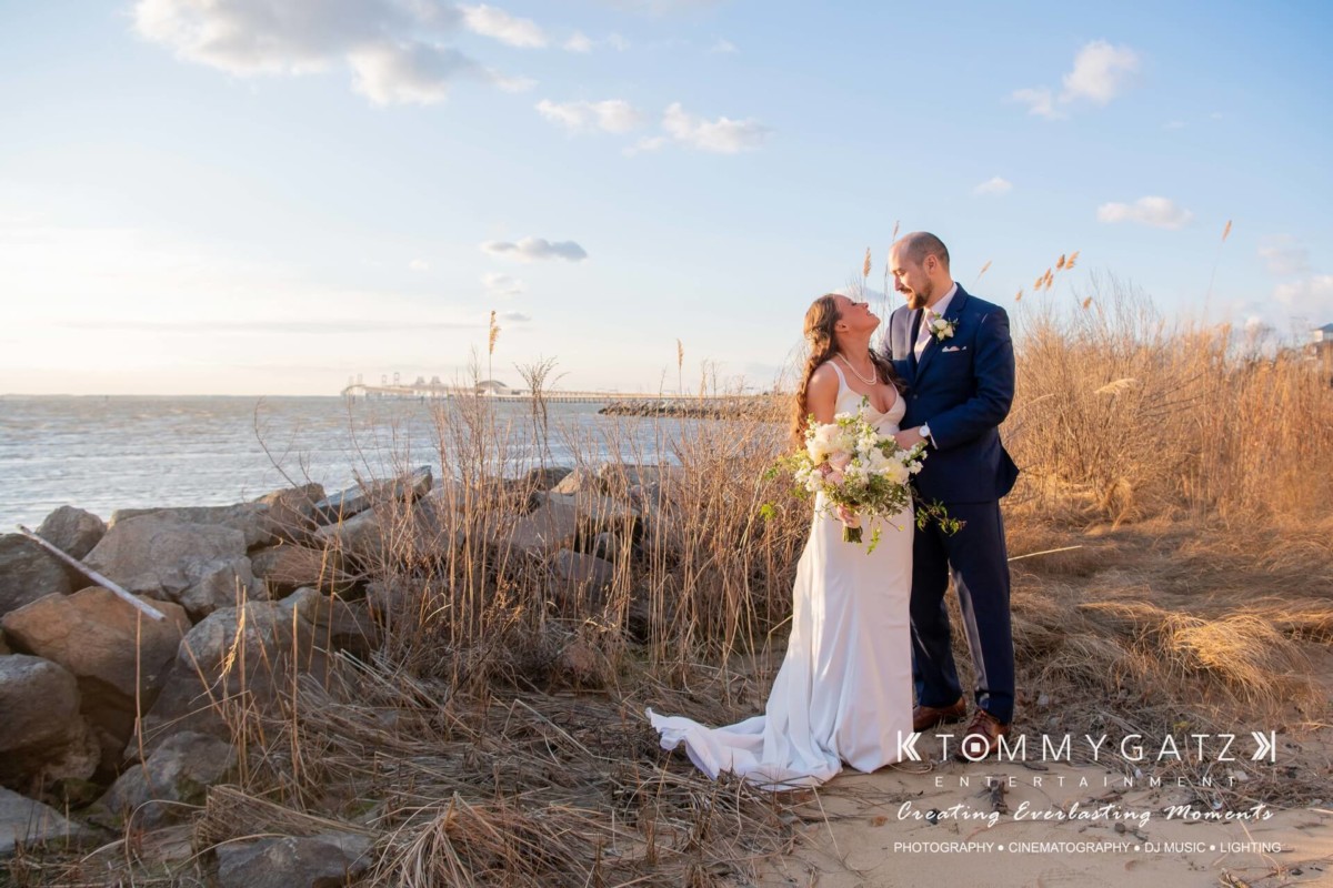 BrittanyDaniel-WEDDING-Highlights-74 - TOMMY GATZ ENTERTAINMENT A couple in wedding attire stands on a grassy shoreline, embracing by the water under a partly cloudy sky.