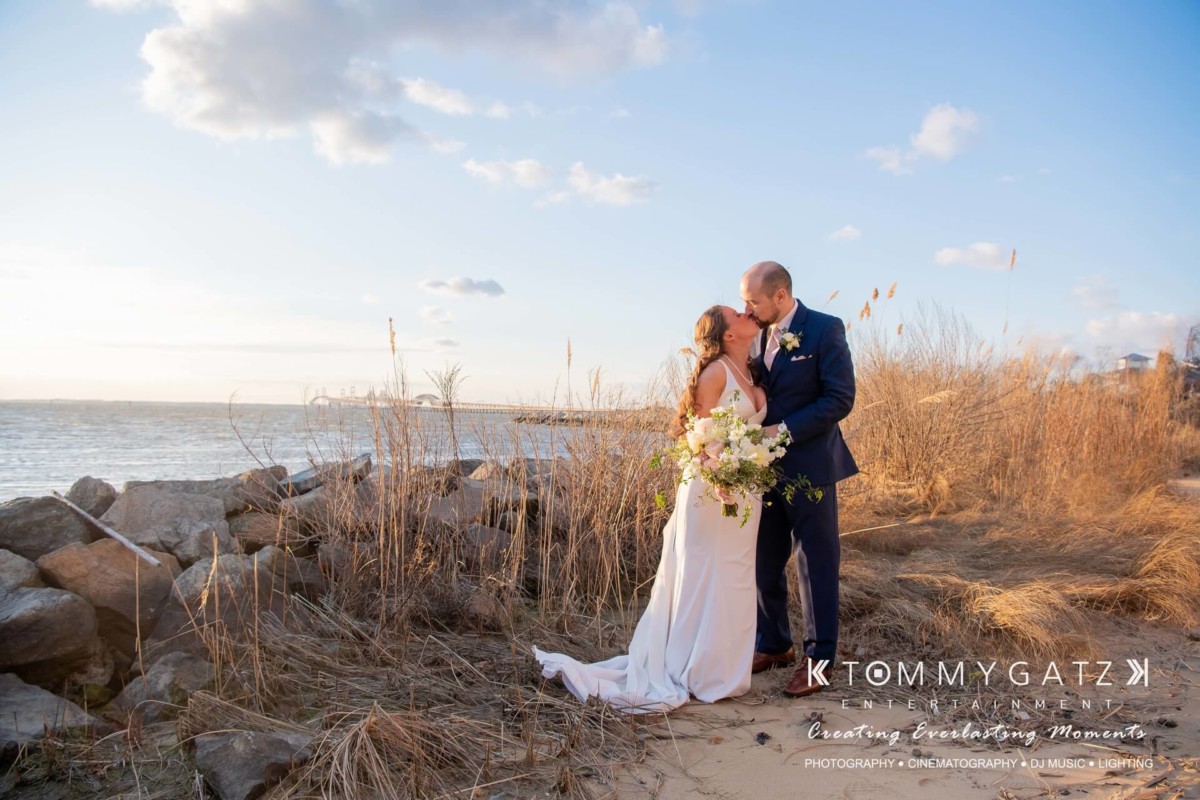 BrittanyDaniel-WEDDING-Highlights-75 - TOMMY GATZ ENTERTAINMENT Wedding couple embracing near a rocky shoreline with tall grass and a calm sea under a clear sky.