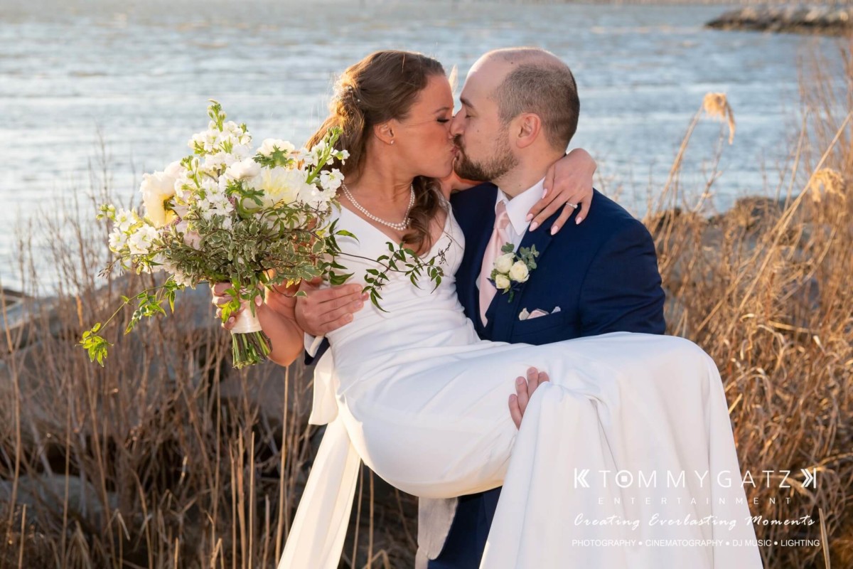 BrittanyDaniel-WEDDING-Highlights-76 - TOMMY GATZ ENTERTAINMENT A bride and groom share a kiss by the water. The groom is wearing a navy suit and carrying the bride, who is holding a bouquet of white flowers.