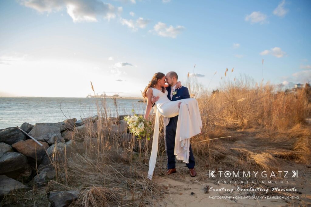 BrittanyDaniel-WEDDING-Highlights-77 - TOMMY GATZ ENTERTAINMENT A couple in wedding attire kiss on a sandy, grassy beach at sunset, with rocks and water in the background.