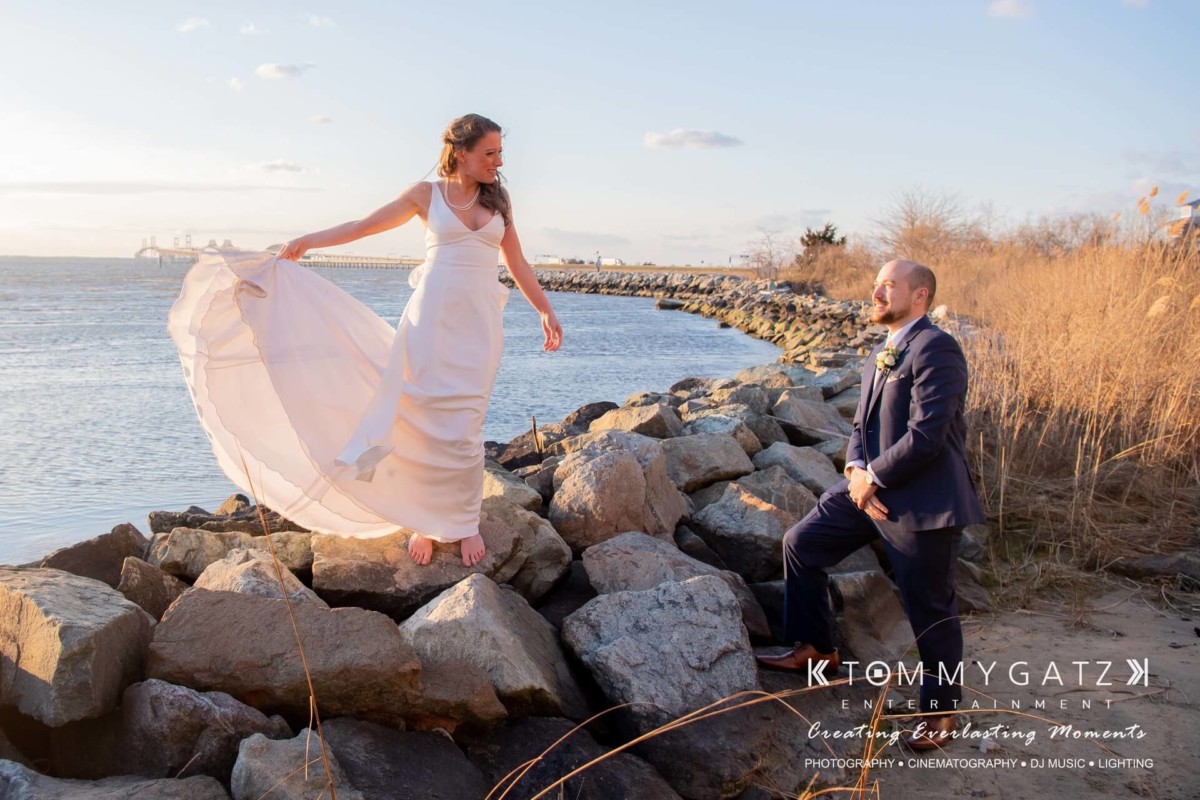 BrittanyDaniel-WEDDING-Highlights-78 - TOMMY GATZ ENTERTAINMENT A bride in a white dress stands barefoot on rocks by the water, while a groom in a suit kneels nearby. It's a sunny day with a clear sky.