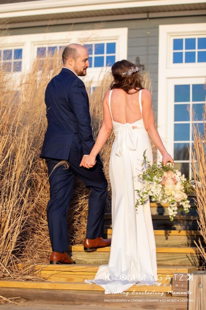 BrittanyDaniel-WEDDING-Highlights-79 - TOMMY GATZ ENTERTAINMENT A bride and groom holding hands, walking up wooden steps outdoors, surrounded by tall grasses. The bride holds a bouquet and wears a white gown; the groom is in a navy suit.