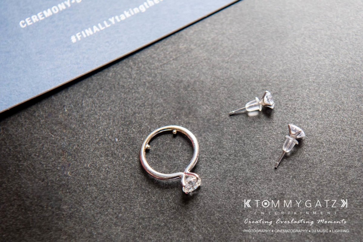 BrittanyDaniel-WEDDING-Highlights-8 - TOMMY GATZ ENTERTAINMENT A diamond ring and a pair of stud earrings are placed on a dark surface near a card with text.