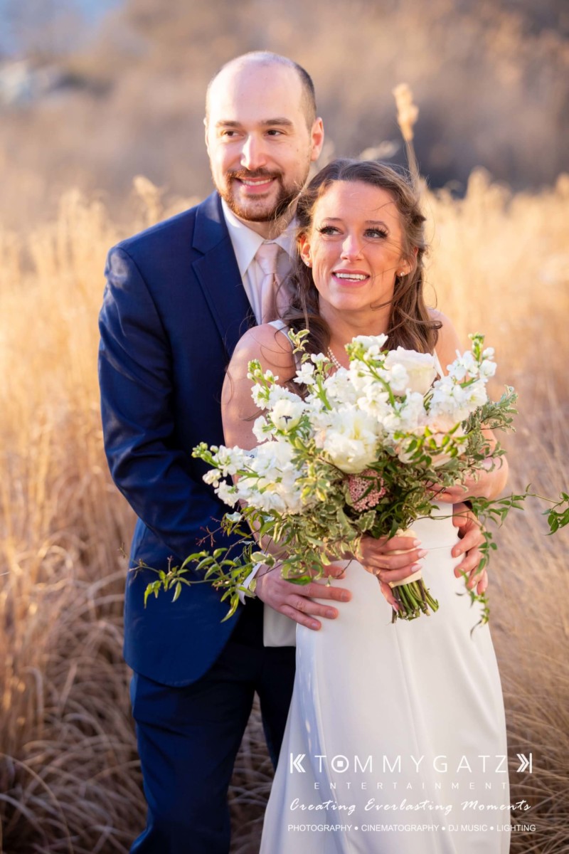 BrittanyDaniel-WEDDING-Highlights-81 - TOMMY GATZ ENTERTAINMENT A couple in formal attire stand in a field. The woman holds a bouquet of white flowers, while the man stands behind her, arms around her waist. They both look toward the distance.