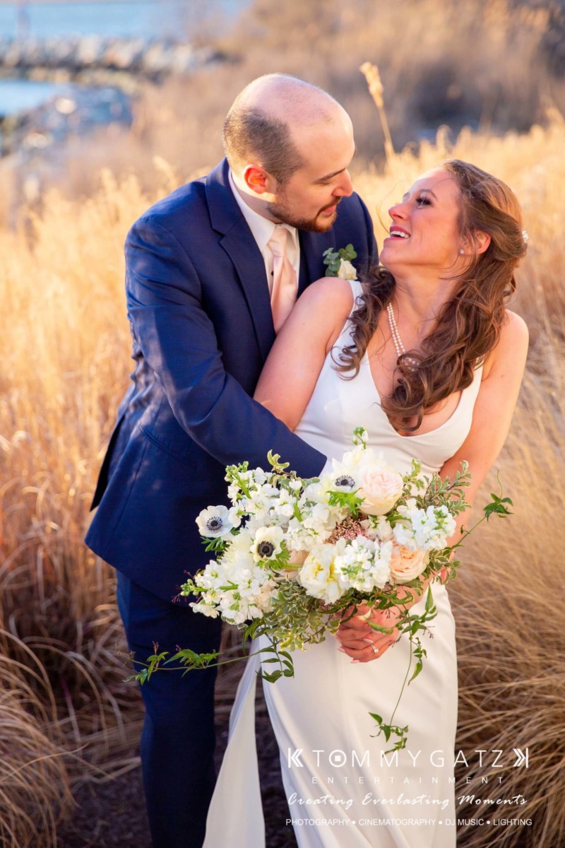 BrittanyDaniel-WEDDING-Highlights-82 - TOMMY GATZ ENTERTAINMENT Bride and groom smiling at each other, standing in front of tall grass, with the bride holding a bouquet of white flowers.
