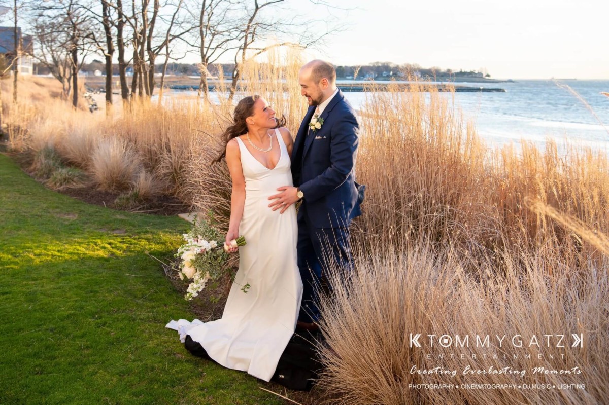 BrittanyDaniel-WEDDING-Highlights-83 - TOMMY GATZ ENTERTAINMENT A bride and groom stand smiling by a lakeside, surrounded by tall grass. The bride holds a bouquet in one hand and her gown's train in the other as the groom rests his hand on her waist.