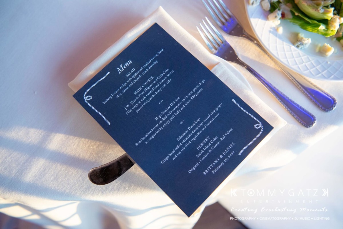 BrittanyDaniel-WEDDING-Highlights-84 - TOMMY GATZ ENTERTAINMENT Dark blue menu on a white plate with silverware, set beside a plate of salad and white flowers.