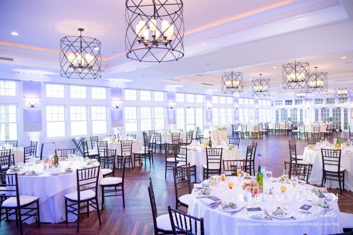 BrittanyDaniel-WEDDING-Highlights-85 - TOMMY GATZ ENTERTAINMENT Elegant banquet hall with round tables set for an event. White tablecloths, wooden chairs, and chandeliers create a formal atmosphere. Large windows line the walls, allowing natural light in.