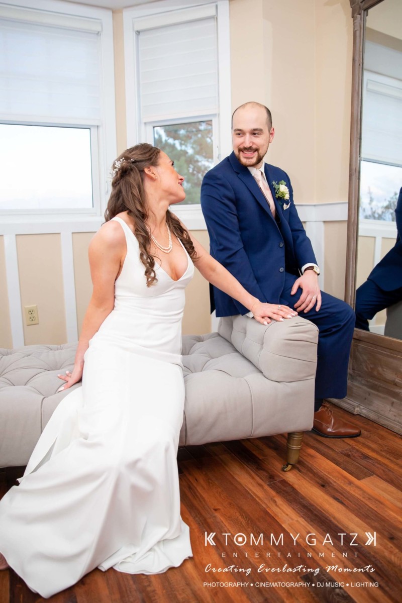 BrittanyDaniel-WEDDING-Highlights-86 - TOMMY GATZ ENTERTAINMENT A bride in a white dress and a man in a suit sit on an upholstered bench, smiling at each other near a large mirror.
