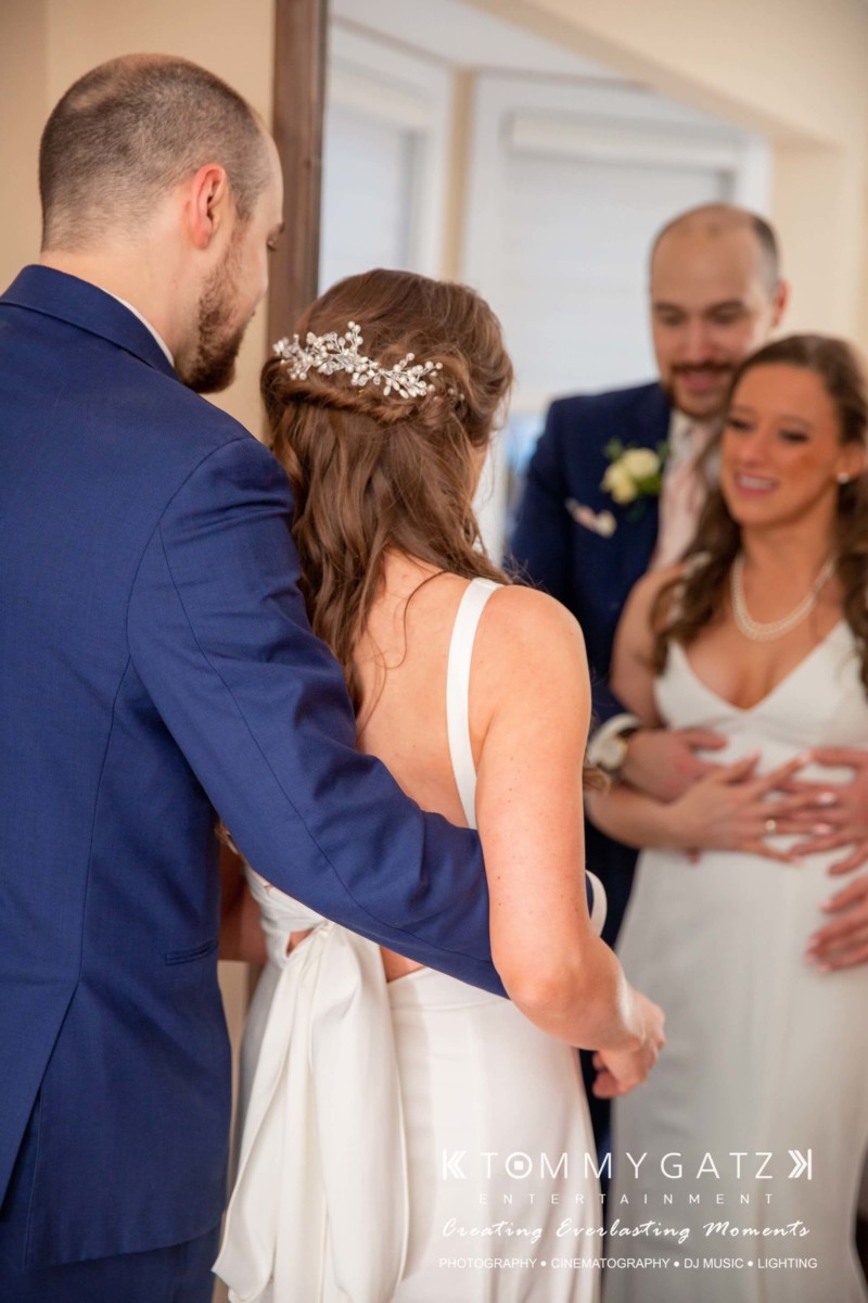 BrittanyDaniel-WEDDING-Highlights-88 - TOMMY GATZ ENTERTAINMENT A bride and groom stand closely together in front of a mirror. Another man in a suit stands nearby, partially visible. The bride wears a white dress with a decorative hairpiece.