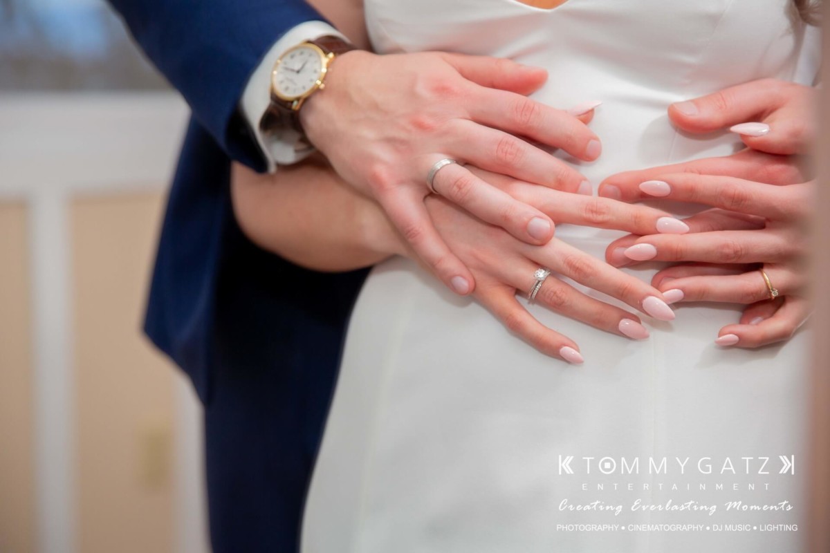 BrittanyDaniel-WEDDING-Highlights-89 - TOMMY GATZ ENTERTAINMENT Close-up of a couple embracing, showing hands with wedding rings. The man's hand supports the woman's waist, over a white dress, showcasing a watch and a suit sleeve.
