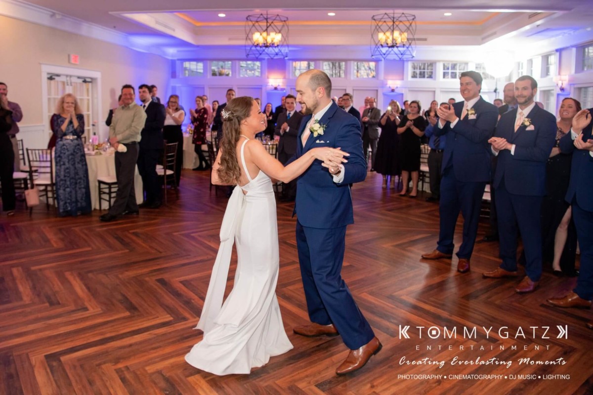BrittanyDaniel-WEDDING-Highlights-92 - TOMMY GATZ ENTERTAINMENT A bride and groom dance in a reception hall as guests look on and clap. The groom is in a blue suit, and the bride in a white dress.