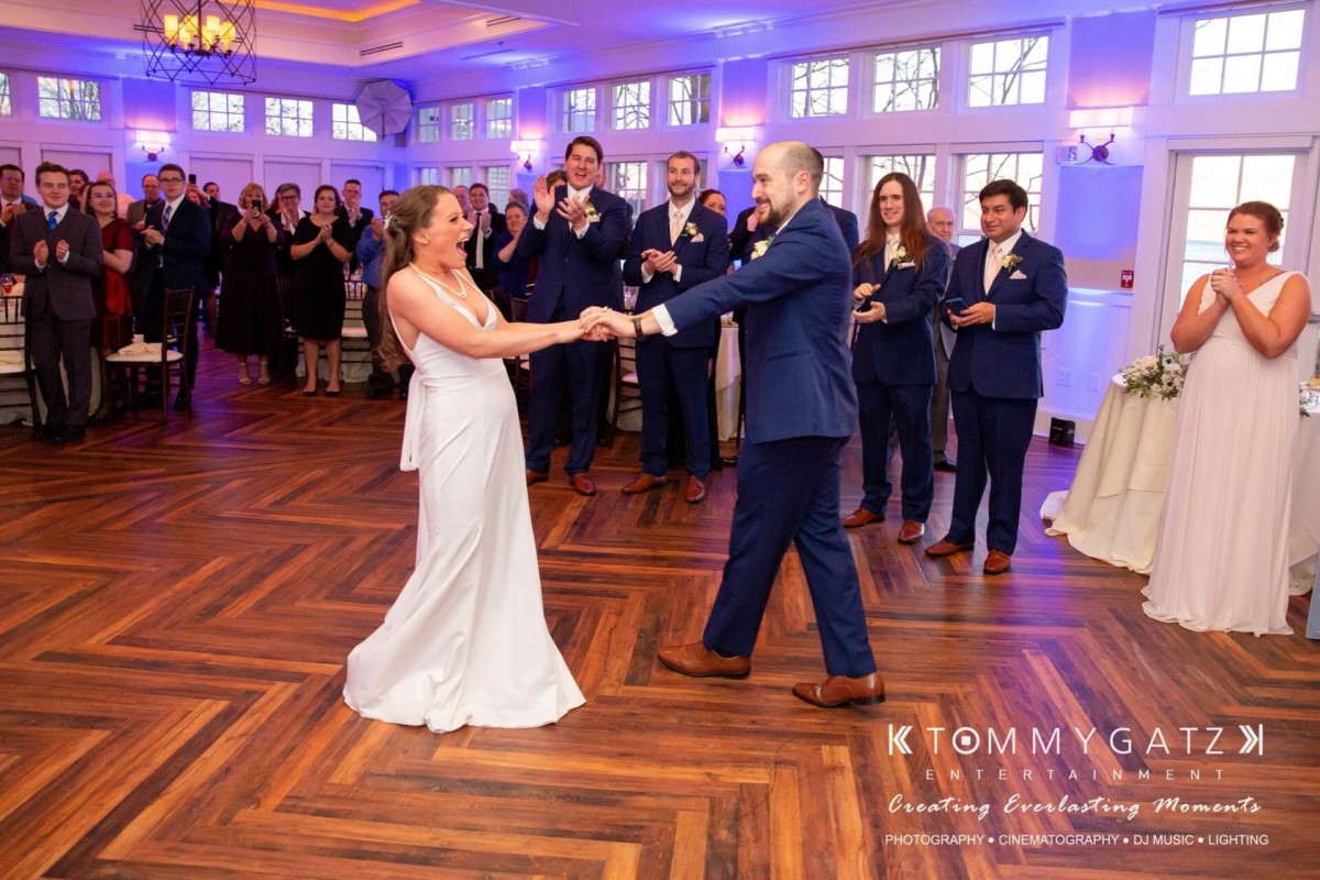 BrittanyDaniel-WEDDING-Highlights-93 - TOMMY GATZ ENTERTAINMENT A bride and groom dance in a room as guests applaud. Elegant decor and wooden floor are visible, with large windows in the background.