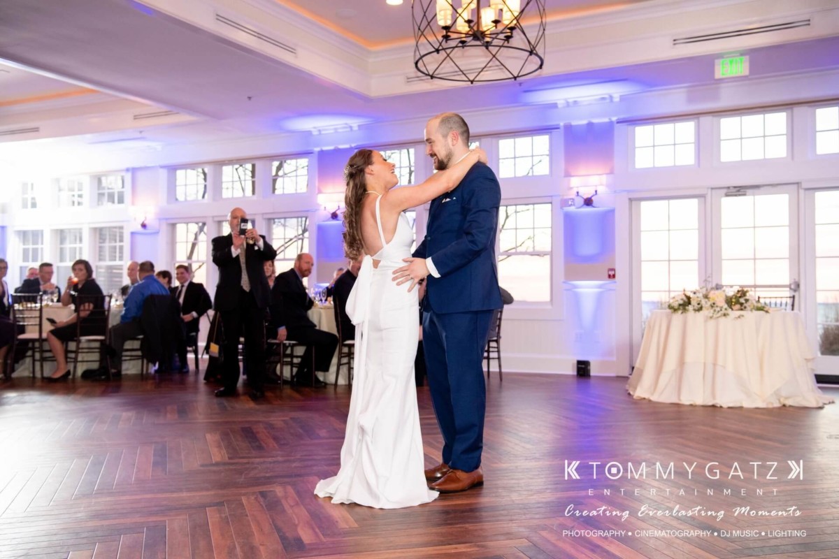 BrittanyDaniel-WEDDING-Highlights-94 - TOMMY GATZ ENTERTAINMENT A bride and groom share their first dance on a wooden floor in a bright reception hall, with seated guests in the background.