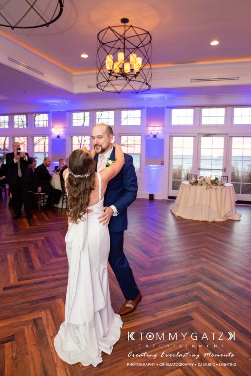 BrittanyDaniel-WEDDING-Highlights-95 - TOMMY GATZ ENTERTAINMENT A couple dances in a decorated ballroom. The woman is in a white dress, and the man is in a suit. Guests are seated at tables in the background.