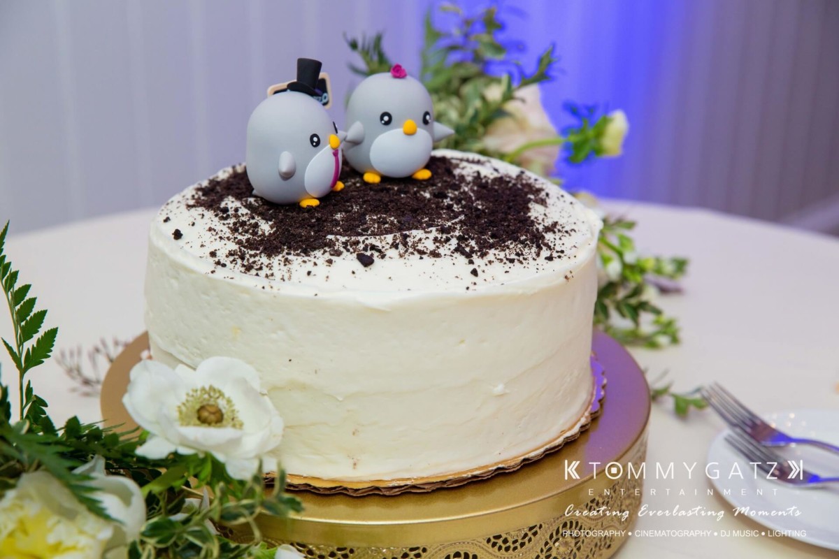 BrittanyDaniel-WEDDING-Highlights-98 - TOMMY GATZ ENTERTAINMENT A white cake topped with two penguin figurines, one wearing a top hat, on a table with flowers and a cake server.