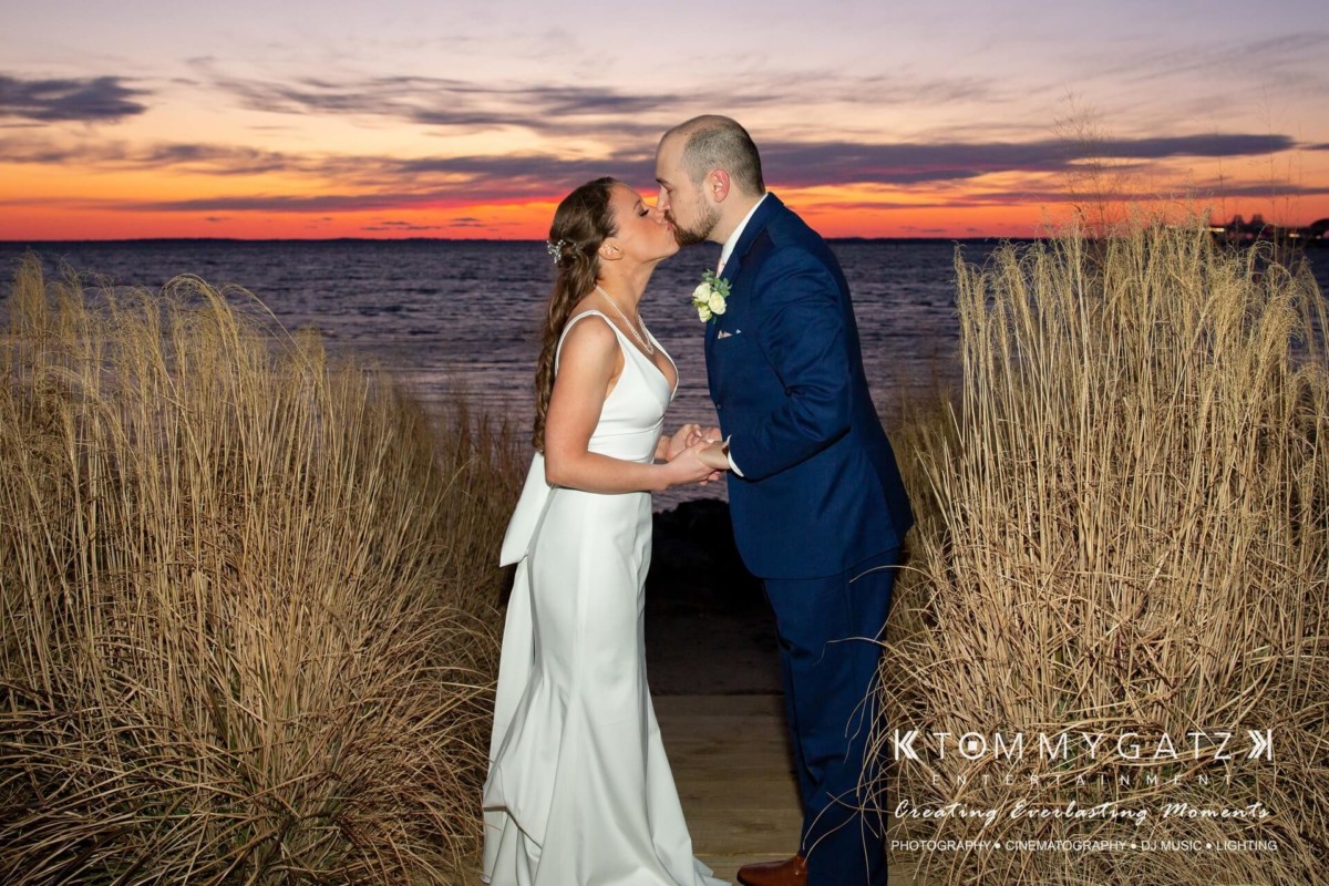 BrittanyDaniel-WEDDING-Highlights-99 - TOMMY GATZ ENTERTAINMENT A bride and groom kiss at a beach during sunset, surrounded by tall grasses.