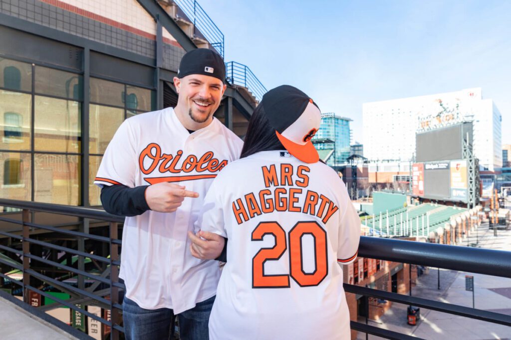 Camden-Yards-Engagment-1 - TOMMY GATZ ENTERTAINMENT A man in an Orioles jersey points at a jersey worn by a person with the name "Mrs. Haggerty" and number 20, standing at a baseball stadium.