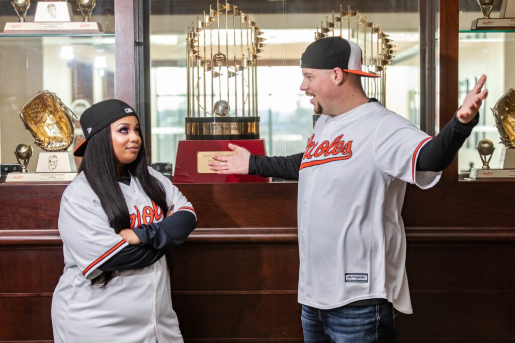 Camden-Yards-Engagment-10 - TOMMY GATZ ENTERTAINMENT Two people in matching baseball jerseys stand in front of a trophy display case. One person gestures widely, while the other crosses their arms.