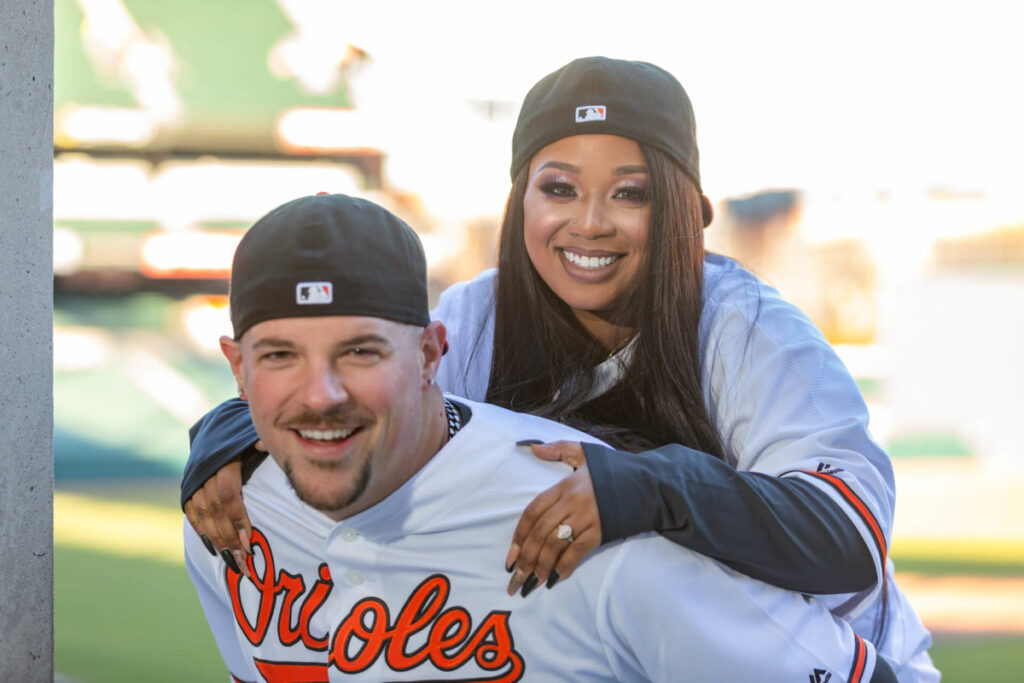 Camden-Yards-Engagment-11 - TOMMY GATZ ENTERTAINMENT Two people wearing baseball jerseys and hats are smiling, with one person playfully on the other's back in a stadium setting.