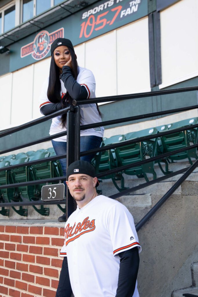 Camden-Yards-Engagment-12 - TOMMY GATZ ENTERTAINMENT Two people in Orioles jerseys, one leaning on a railing and the other standing on stairs, outside a sports venue.