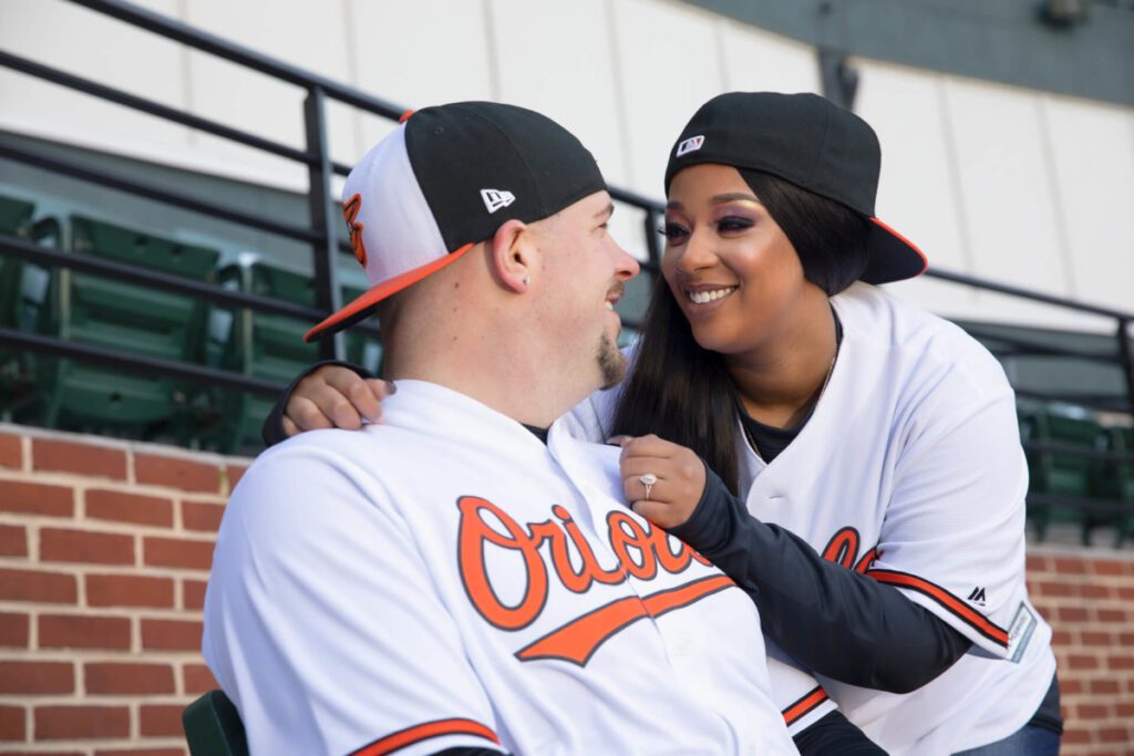 Camden-Yards-Engagment-13 - TOMMY GATZ ENTERTAINMENT Two people in matching Orioles jerseys and caps, sitting together in a baseball stadium, smiling at each other.