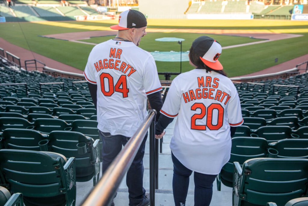 Camden-Yards-Engagment-14 - TOMMY GATZ ENTERTAINMENT A couple wearing matching jerseys labeled "Mr. Haggerty 04" and "Mrs. Haggerty 20" holds hands in an empty baseball stadium.