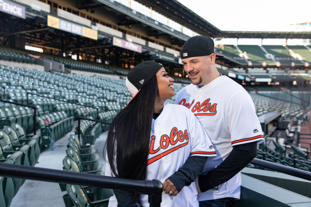 Camden-Yards-Engagment-16 - TOMMY GATZ ENTERTAINMENT A couple wearing Orioles jerseys and caps smile at each other while standing in the stadium seating area.
