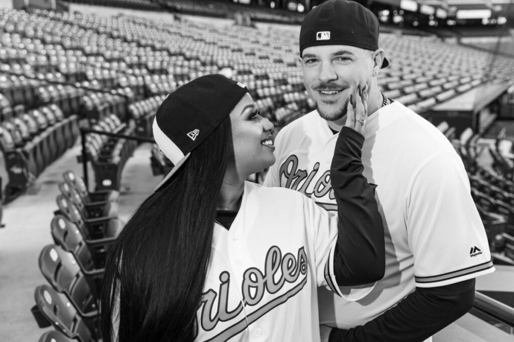 Camden-Yards-Engagment-17 - TOMMY GATZ ENTERTAINMENT Two people wearing Orioles baseball jerseys and caps stand in an empty stadium. One smiles while touching the other's face.