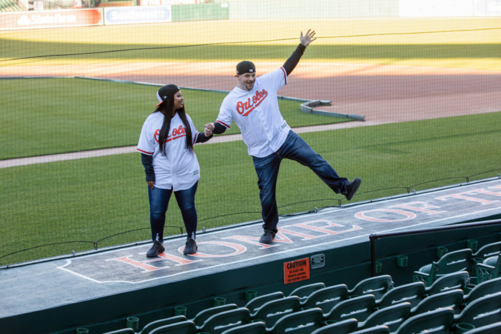 Camden-Yards-Engagment-18 - TOMMY GATZ ENTERTAINMENT Two people wearing Orioles baseball jerseys stand on the dugout edge at a stadium, with one playfully posing while the other looks on.