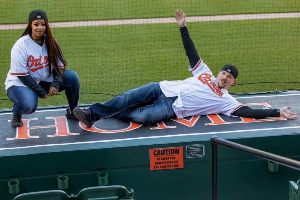 Camden-Yards-Engagment-19 - TOMMY GATZ ENTERTAINMENT Two people in Orioles jerseys pose on the "HOME" dugout bench at a baseball field. One kneels, and the other is lying down with arms outstretched, both smiling.