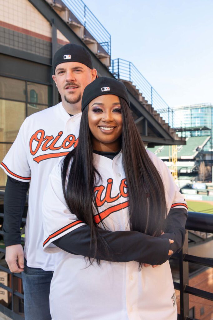 Camden-Yards-Engagment-2 - TOMMY GATZ ENTERTAINMENT Two people wearing baseball jerseys and caps pose in a stadium. The woman is in front, smiling, while the man stands behind her.