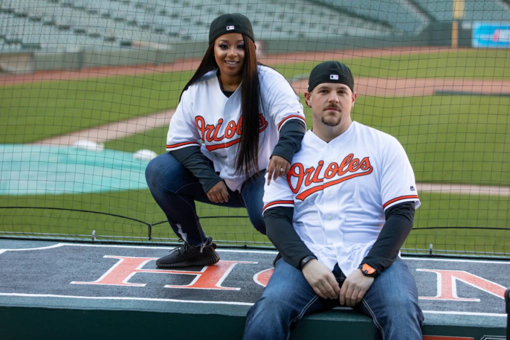 Camden-Yards-Engagment-21 - TOMMY GATZ ENTERTAINMENT Two people wearing Orioles baseball jerseys and caps pose in a stadium, sitting near the edge of a platform with the field visible in the background.
