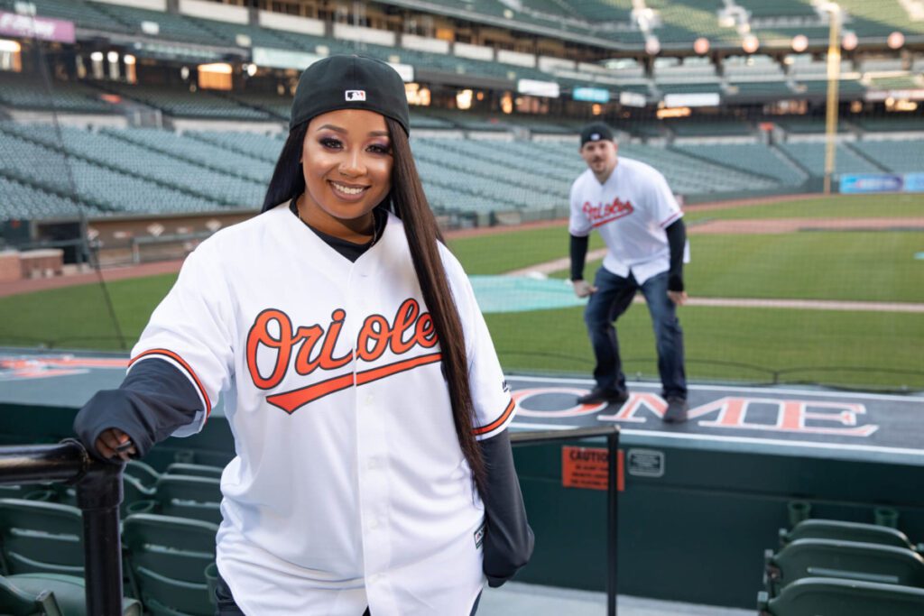 Camden-Yards-Engagment-22 - TOMMY GATZ ENTERTAINMENT Two people wearing Orioles jerseys and backward caps stand on a baseball field, with one posing in the foreground and the other in the background.