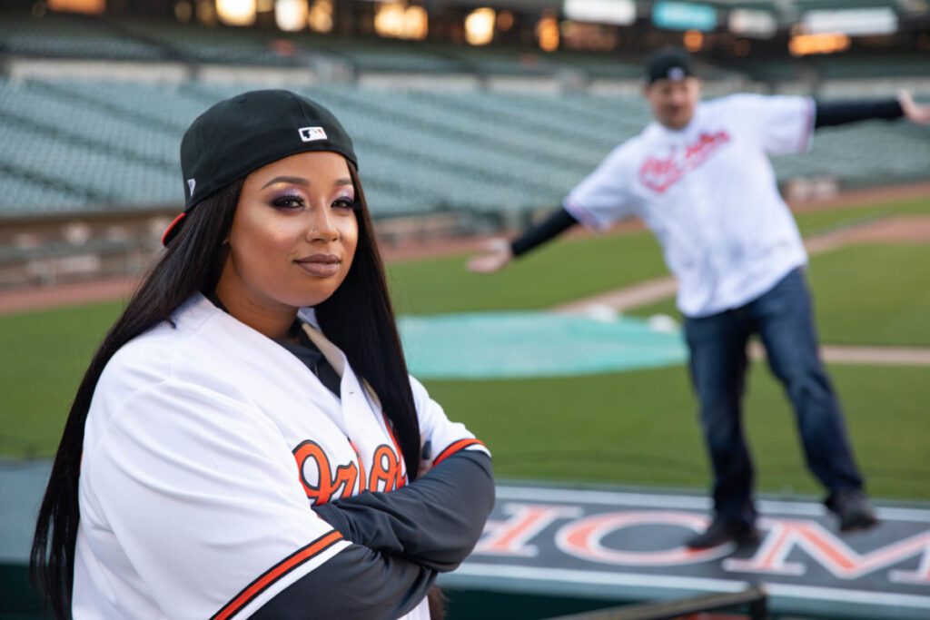 Camden-Yards-Engagment-24 - TOMMY GATZ ENTERTAINMENT Two people in baseball jerseys on a field; one is posing confidently in the foreground, while the other playfully gestures in the background.