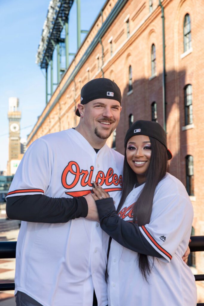 Camden-Yards-Engagment-3 - TOMMY GATZ ENTERTAINMENT A couple in matching baseball jerseys and caps stands smiling in front of a red brick building with a clock tower in the background.