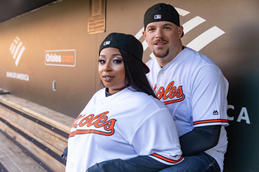 Camden-Yards-Engagment-30 - TOMMY GATZ ENTERTAINMENT Two people wearing Baltimore Orioles baseball jerseys and caps sit together in a dugout.