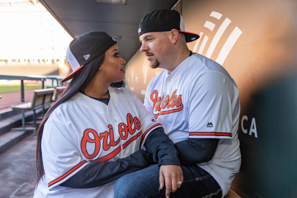 Camden-Yards-Engagment-31 - TOMMY GATZ ENTERTAINMENT Two people wearing Orioles baseball jerseys and caps, sitting and looking at each other in a dugout.