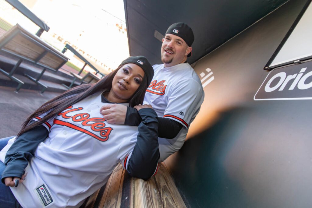 Camden-Yards-Engagment-32 - TOMMY GATZ ENTERTAINMENT Two people in baseball jerseys pose together in a dugout, smiling and leaning on a wooden bench.